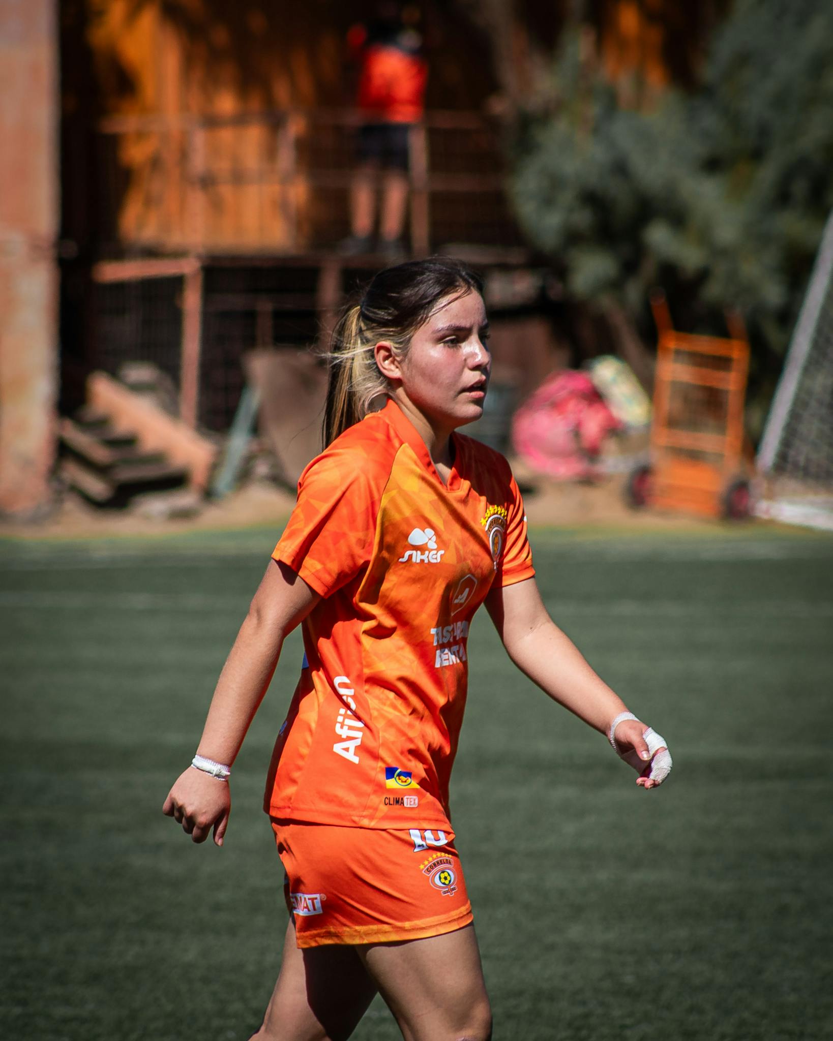 Female soccer player in orange uniform walking on the field during day match.