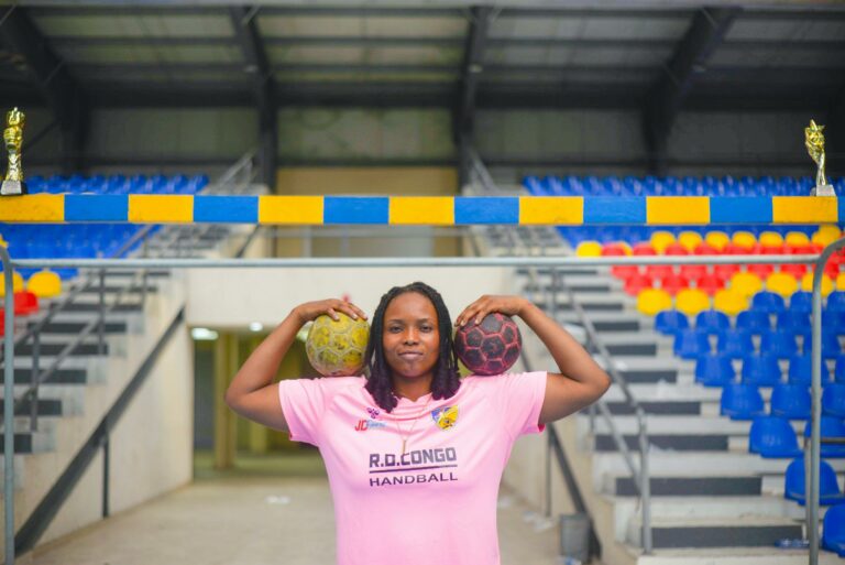Female athlete in a pink handball jersey poses confidently inside a vibrant sports arena with handballs and trophies.