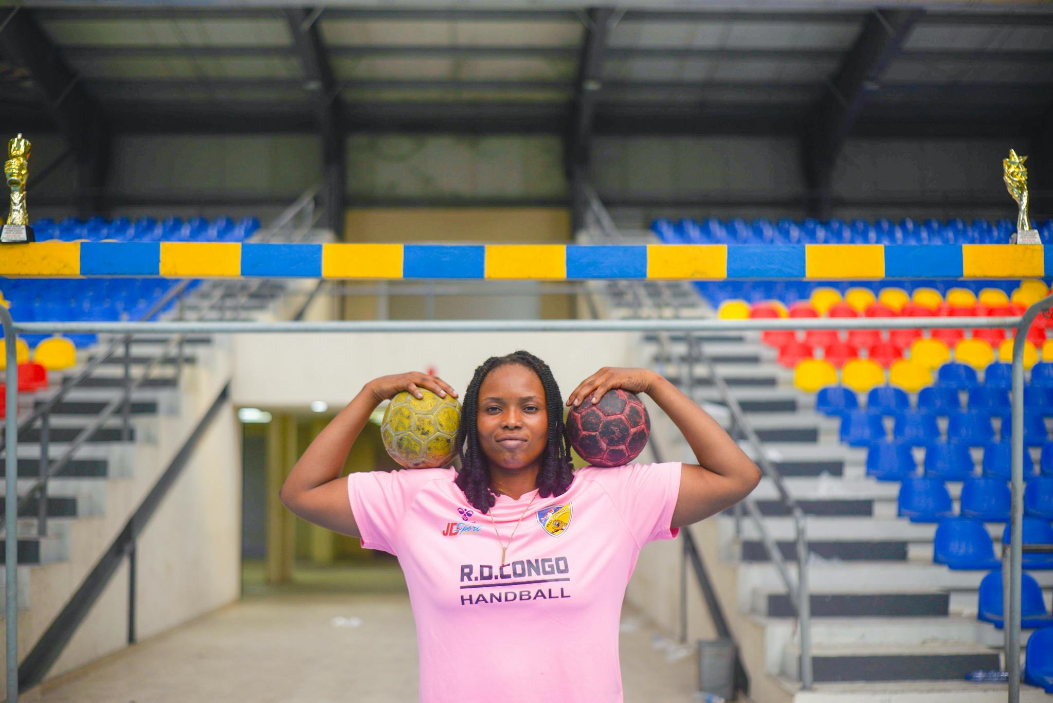 Female athlete in a pink handball jersey poses confidently inside a vibrant sports arena with handballs and trophies.