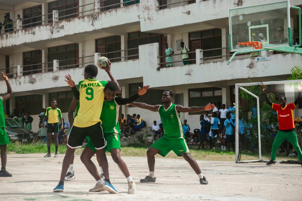 Lively outdoor handball match at a school with players in green and yellow uniforms and vibrant crowd in the background.